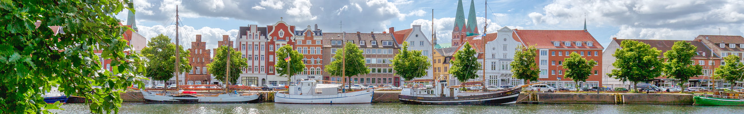 Historische Altstadt Lübecks mit bunten Giebelhäusern, Segelschiffen und der Trave unter blauem Himmel mit Wolken.